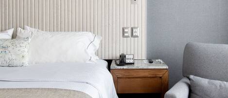 Una habitación de hotel con una cama grande, una mesita de noche con un teléfono y un sillón gris.