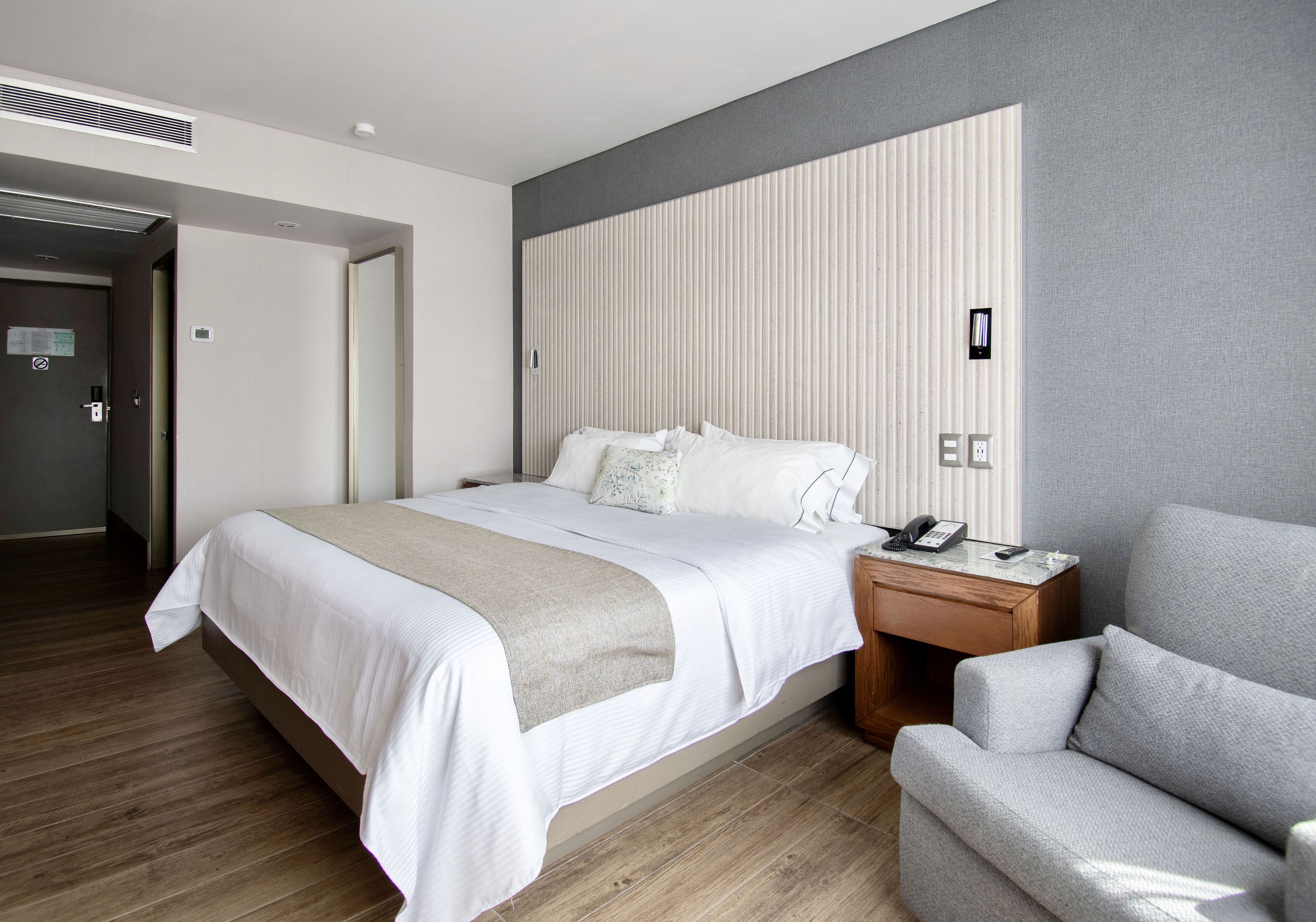 Una habitación de hotel moderna con una cama grande, una mesita de noche con teléfono y lámpara, un sillón gris y suelo de madera.