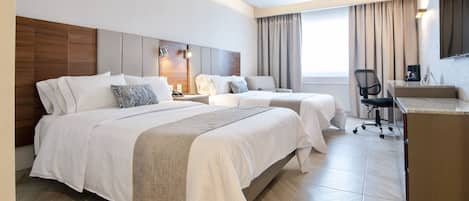 Habitación de hotel con dos camas, un escritorio, una silla y un ventanal grande con cortinas.