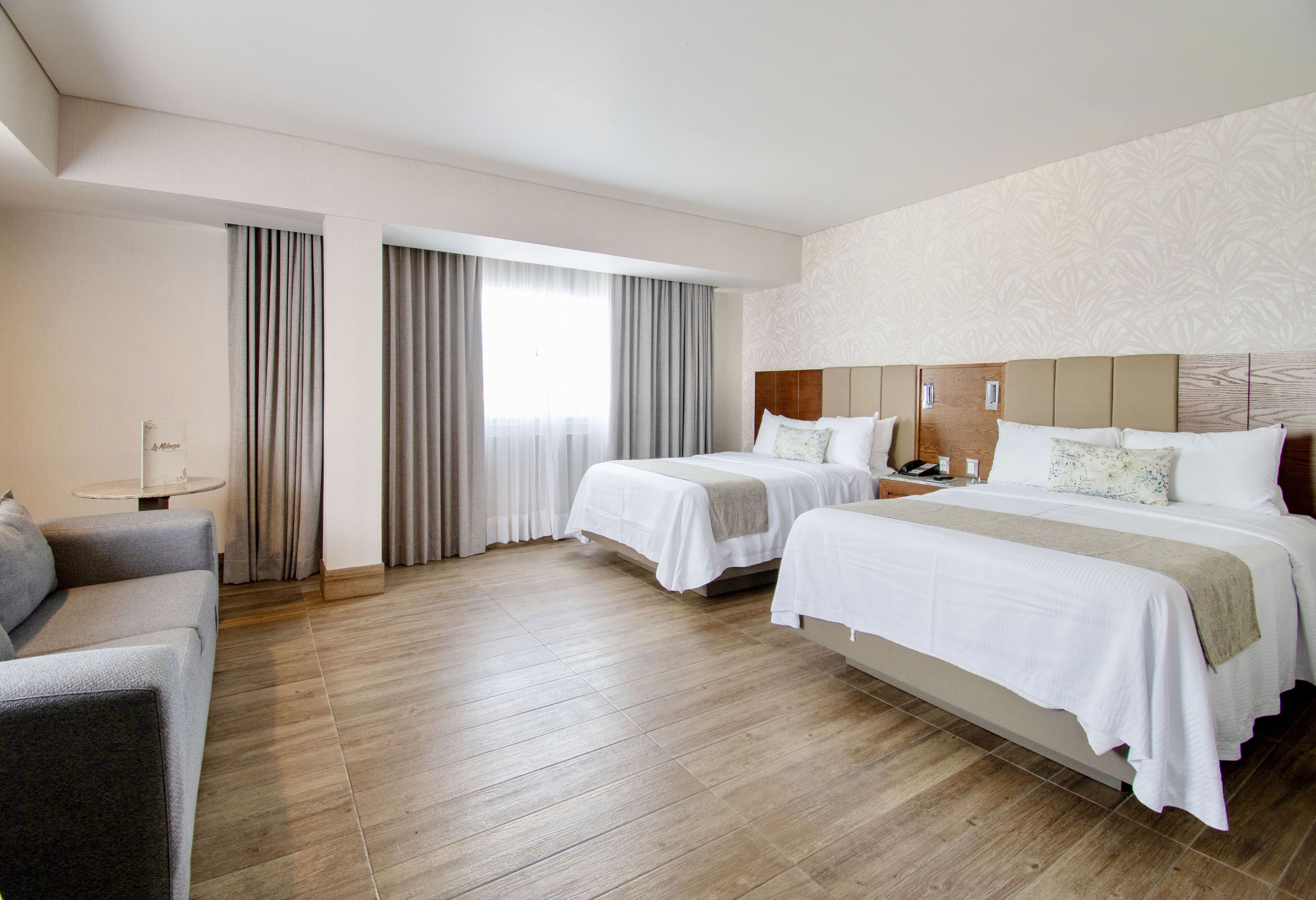 A hotel room with two beds, a sofa, a small table, and a large window with curtains.