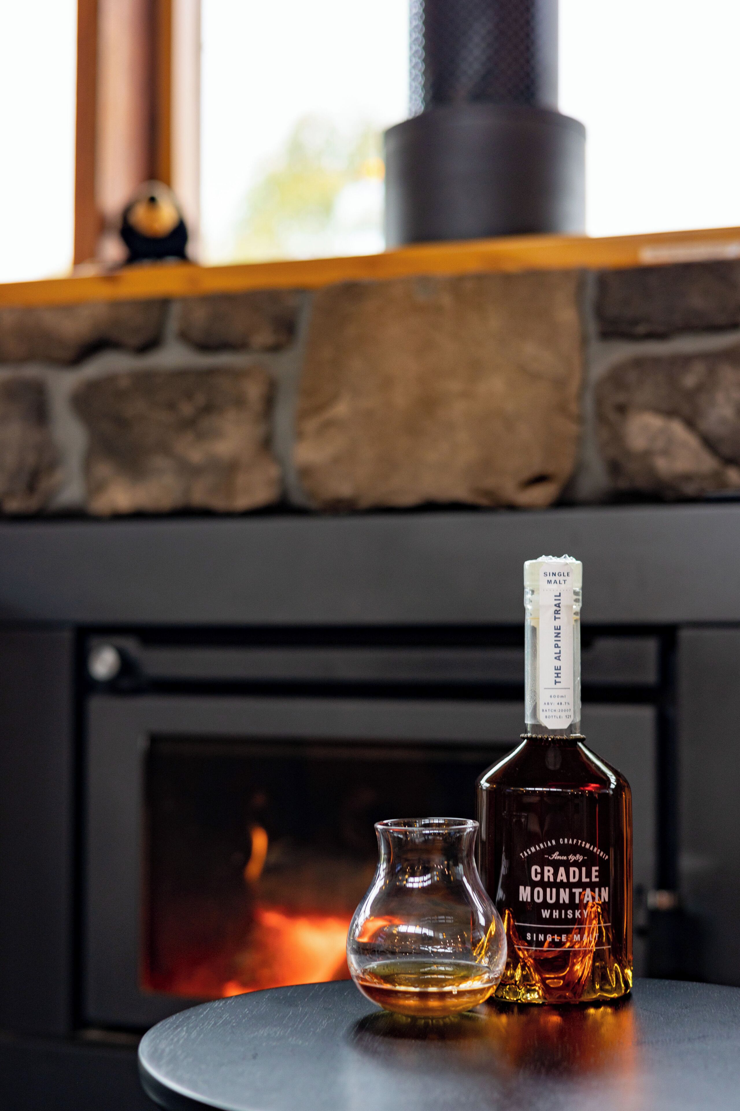 Una bottiglia di whisky Cradle Mountain e un bicchiere su un tavolo di fronte a un camino.