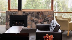 Una sala de estar acogedora con chimenea de piedra, un sofá negro, una mesa de madera y un amplio ventanal con vistas a árboles.