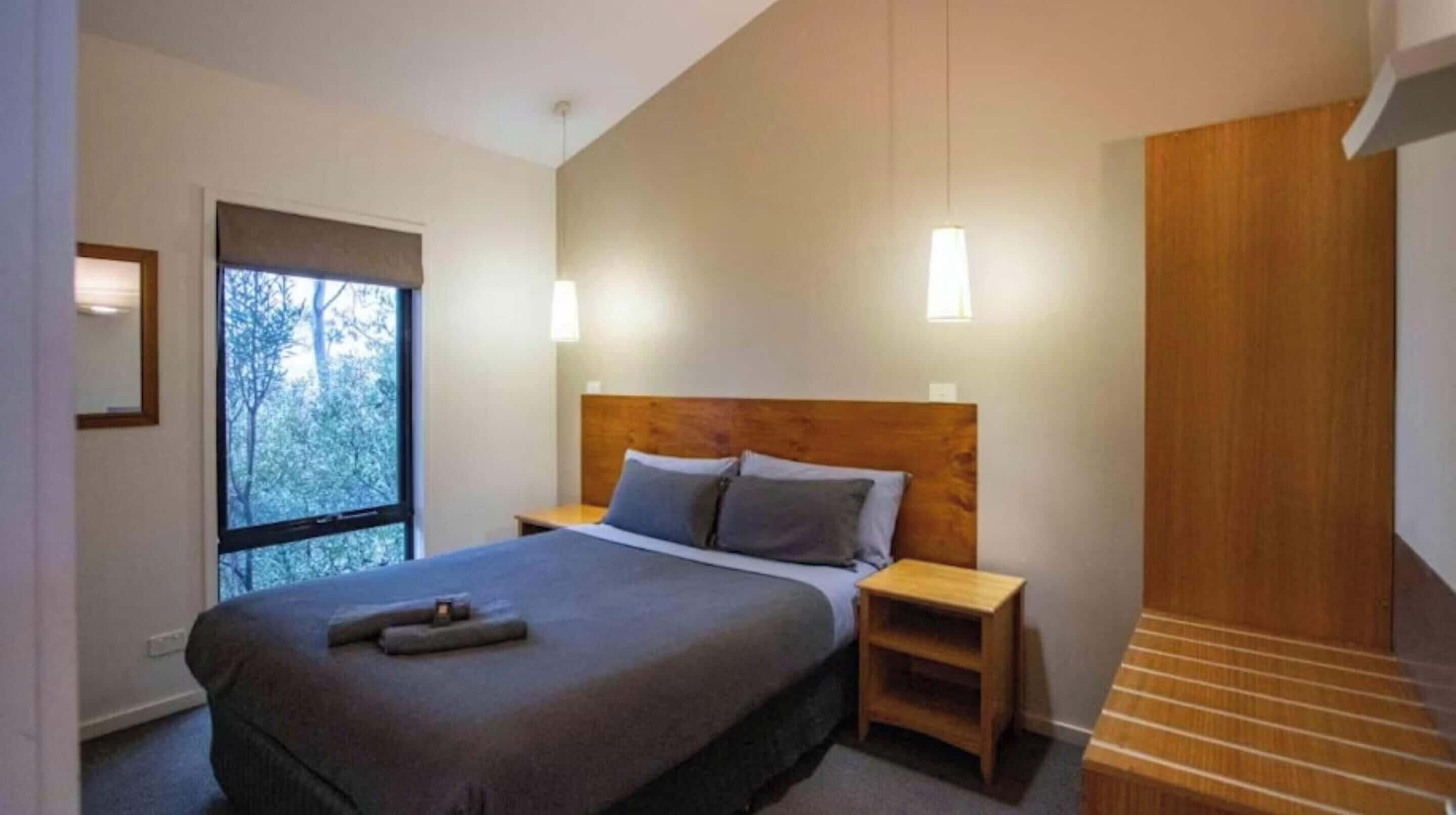 Ein modernes Schlafzimmer mit einem großen Bett, einem Holz-Kopfende, Nachttischen und einem Fenster mit Blick auf Bäume.