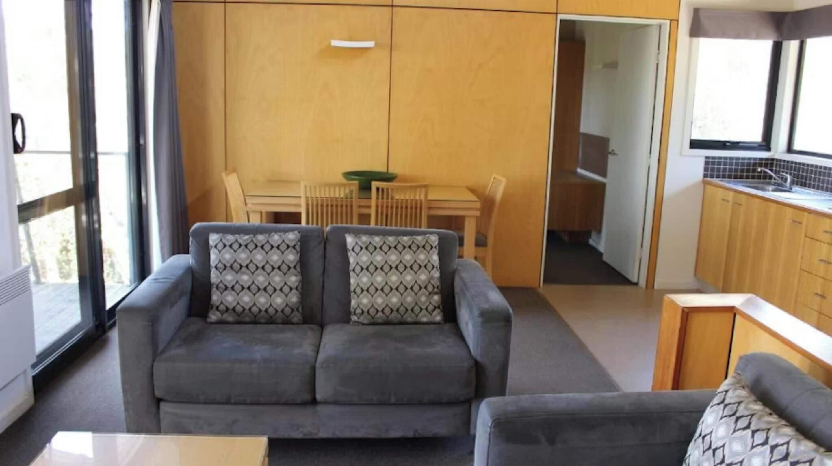 A modern living room with a grey sofa and patterned cushions, a dining area with a wooden table and chairs, and a kitchen with wooden cabinets and a window.