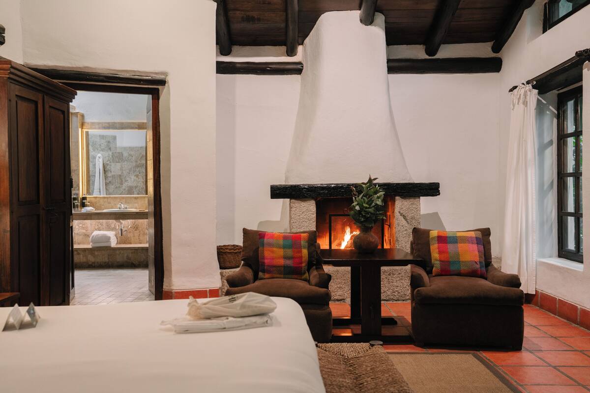 Une chambre à coucher avec un lit, une cheminée, deux fauteuils et une vue sur la salle de bain.