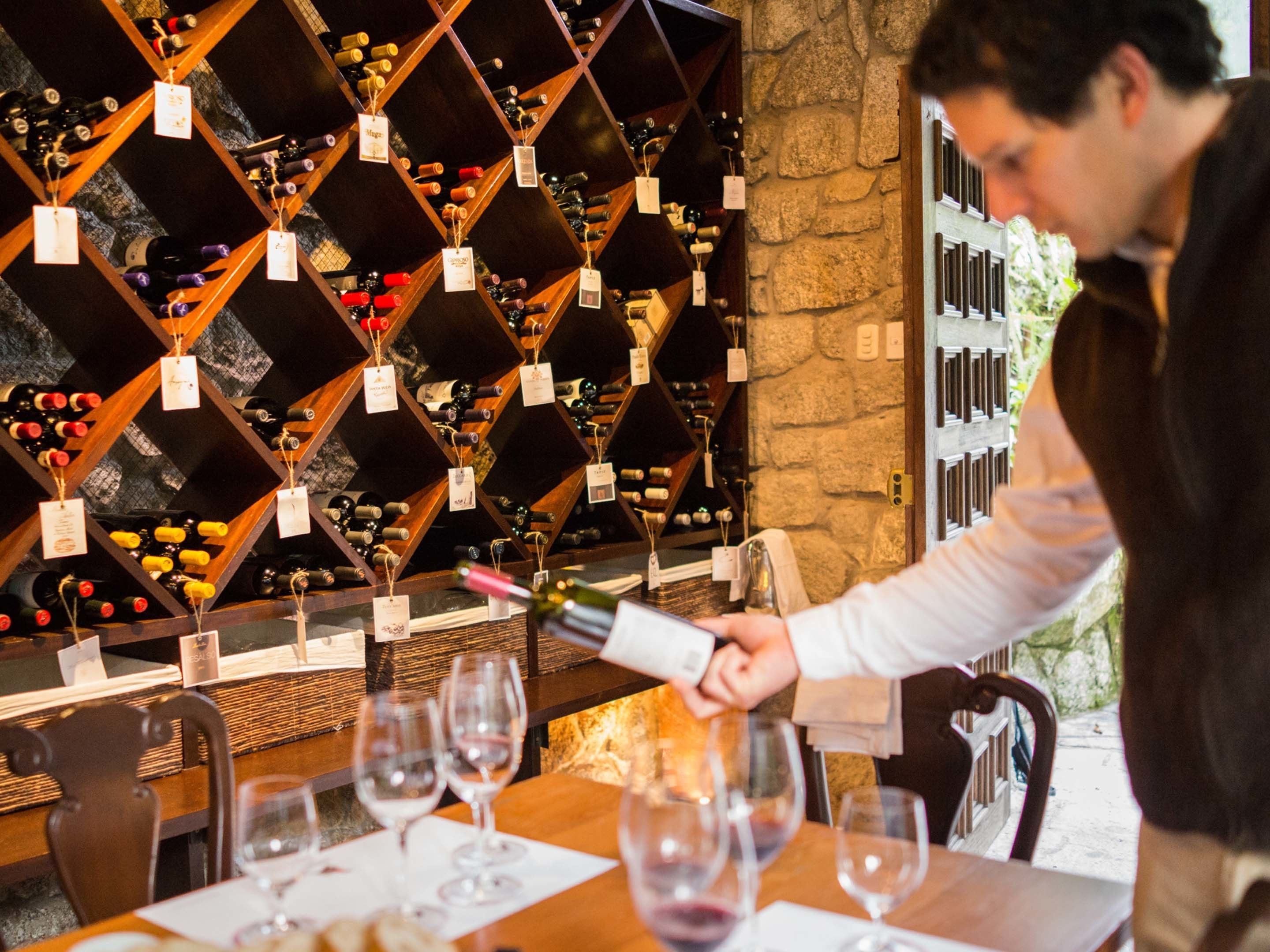 A wine cellar with wooden racks holding bottles, a person pouring wine, and empty wine glasses on a table.