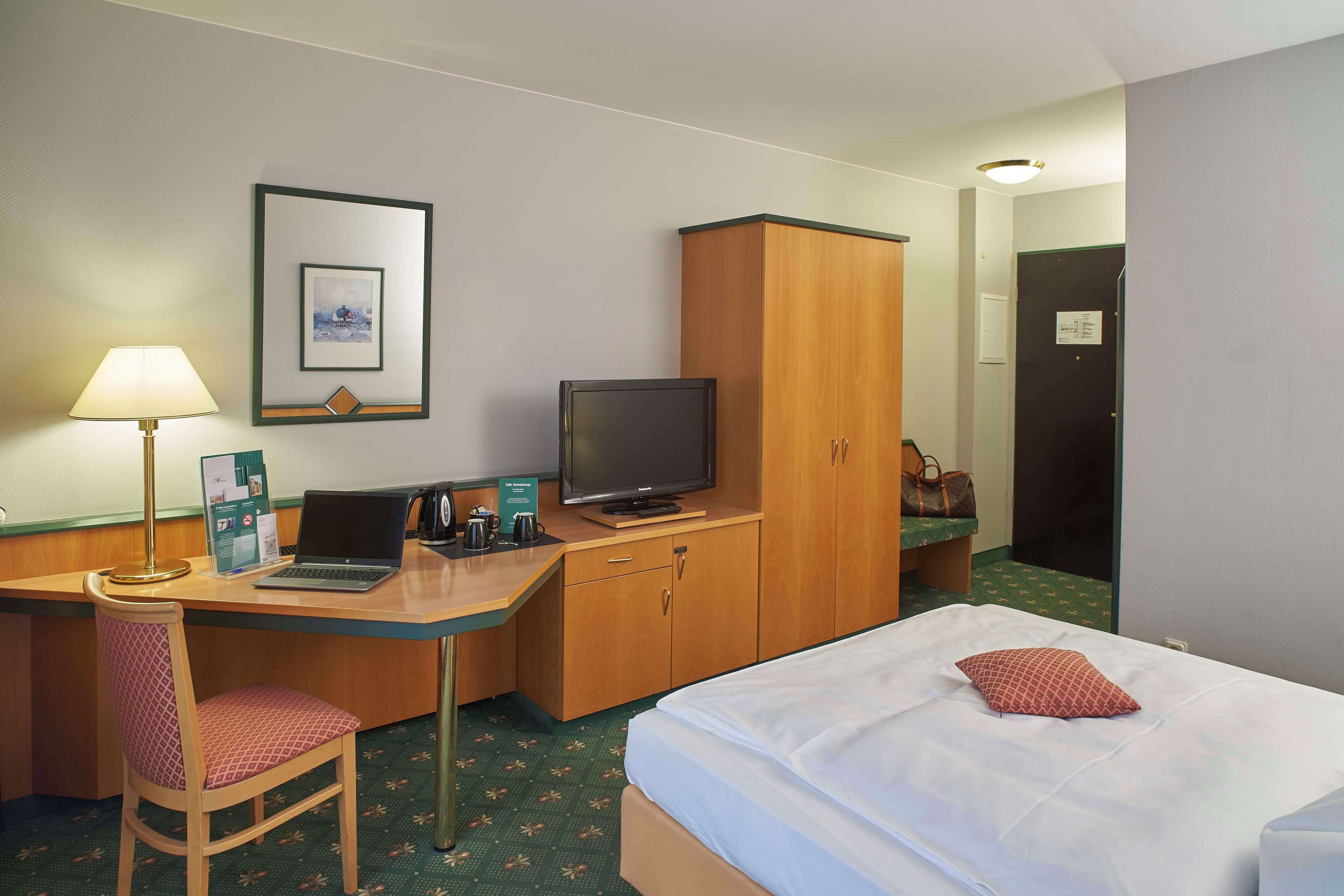 A hotel room with a bed, a desk with a laptop, a television, a chair, a lamp, and a wardrobe.