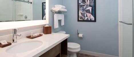A modern bathroom with a large sink, a toilet, and a shower. There is a towel rack with towels, a framed abstract painting, and a small wooden tray on the sink.