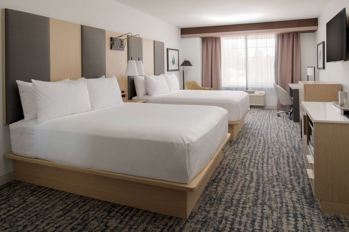 A hotel room with two beds, a desk, a chair, a TV, and a window with curtains.