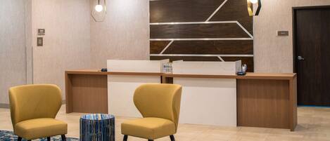 A reception area with a wooden desk, two yellow chairs, a patterned ottoman, and a geometric wall design.