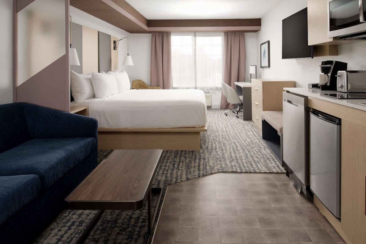 A modern hotel room with a bed, a sofa, a desk, a kitchenette, and a large window.