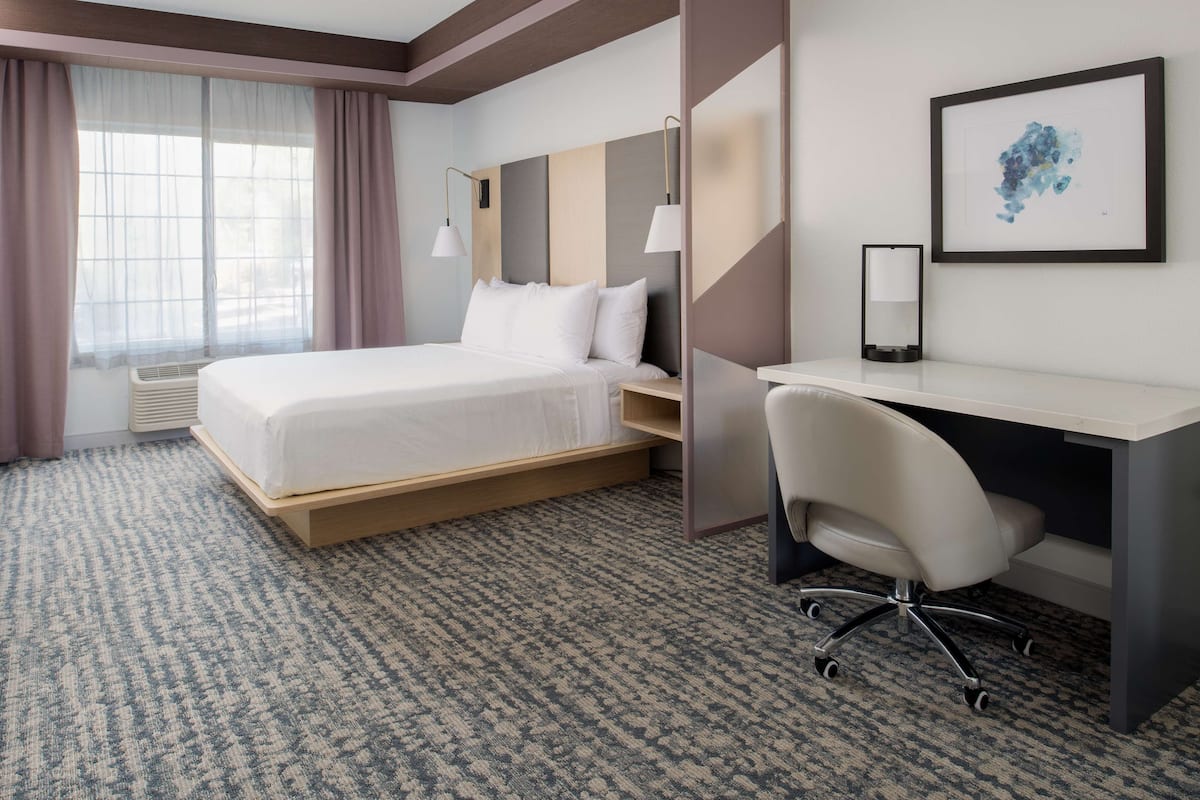 A hotel room with a bed, desk, chair, and a framed artwork on the wall.