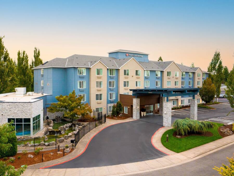 Un hotel de varios pisos con fachada azul y beige, una entrada cubierta y un camino de acceso arbolado.