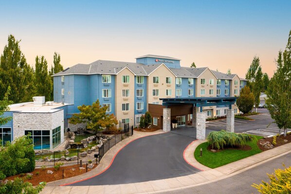 A multi-story hotel with a blue and beige facade, a covered entrance, and a landscaped driveway.