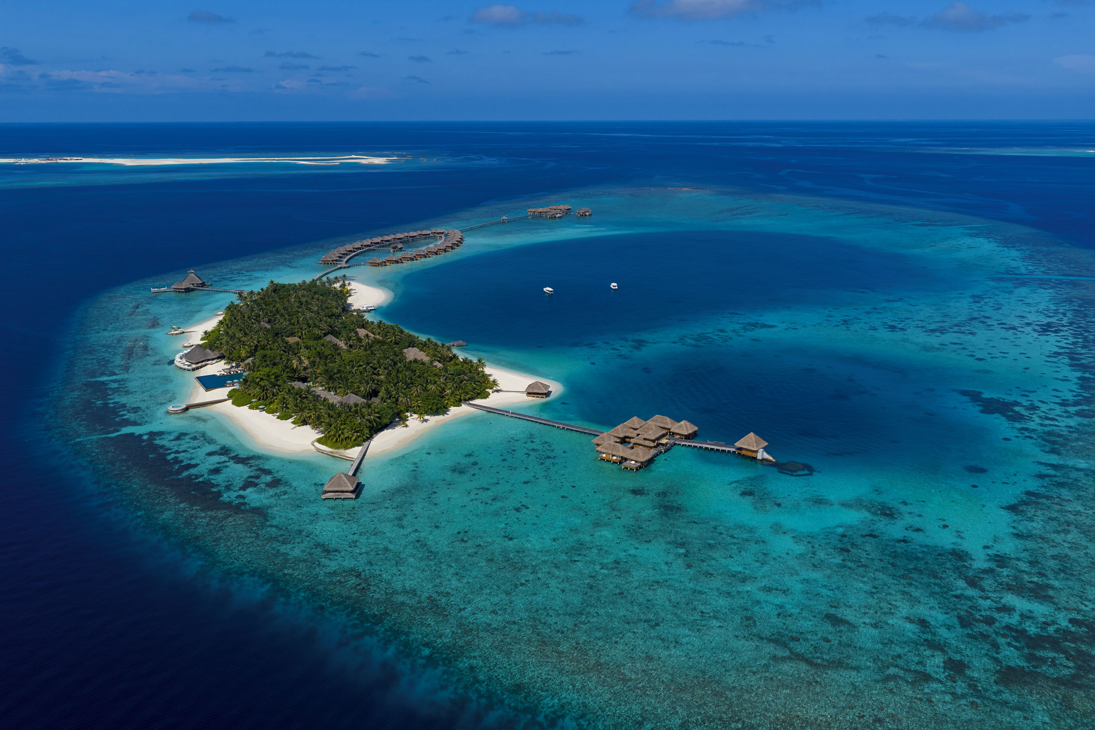 An aerial view of a tropical island resort with overwater bungalows, clear turquoise waters, and a small sandy beach.