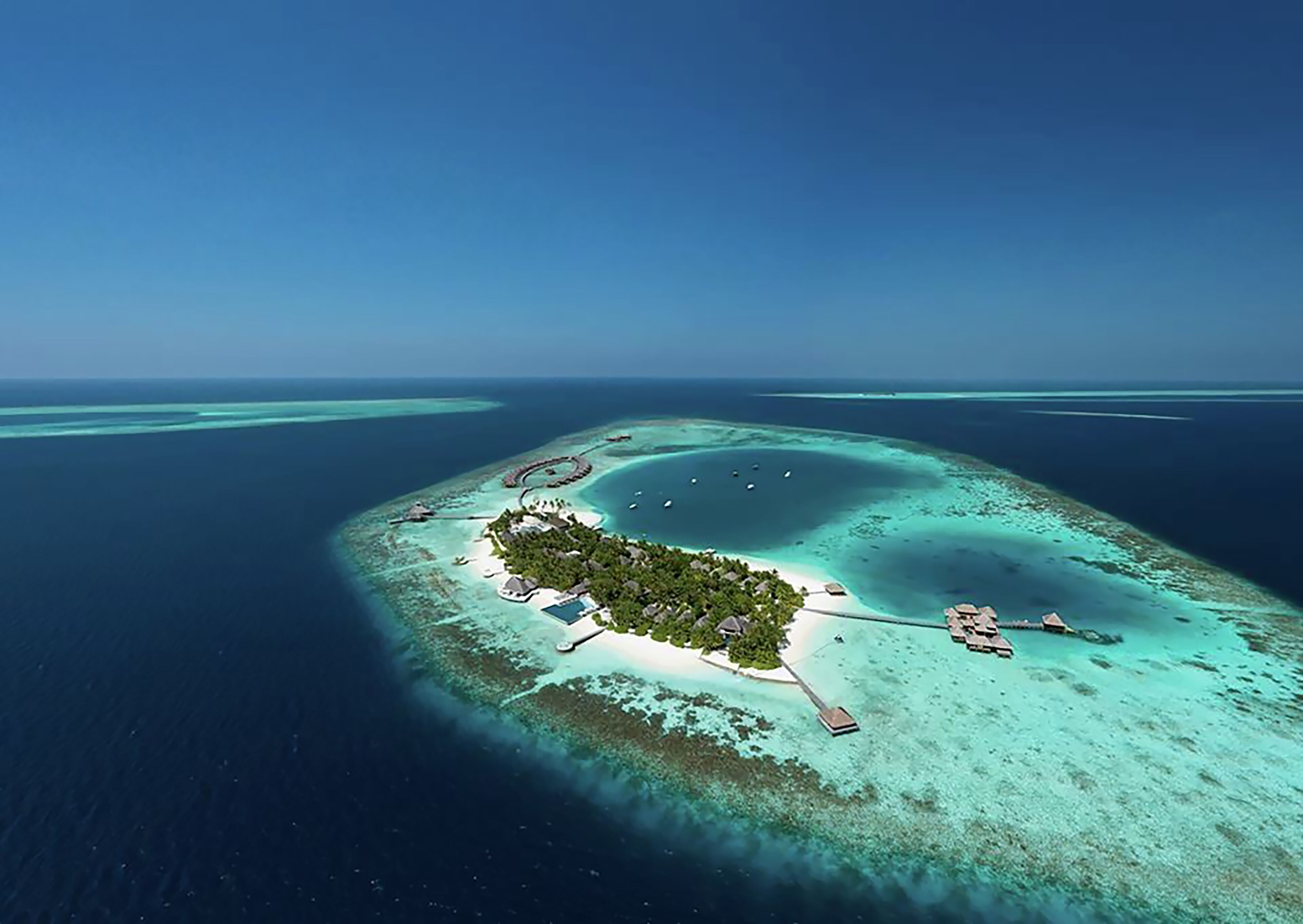 An aerial view of a tropical island with clear turquoise waters, a central lagoon, and several overwater bungalows.