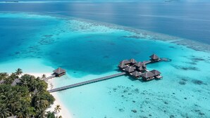 Luchtbeeld van overwater bungalows verbonden door een brug, omgeven door turquoise water en palmbomen.