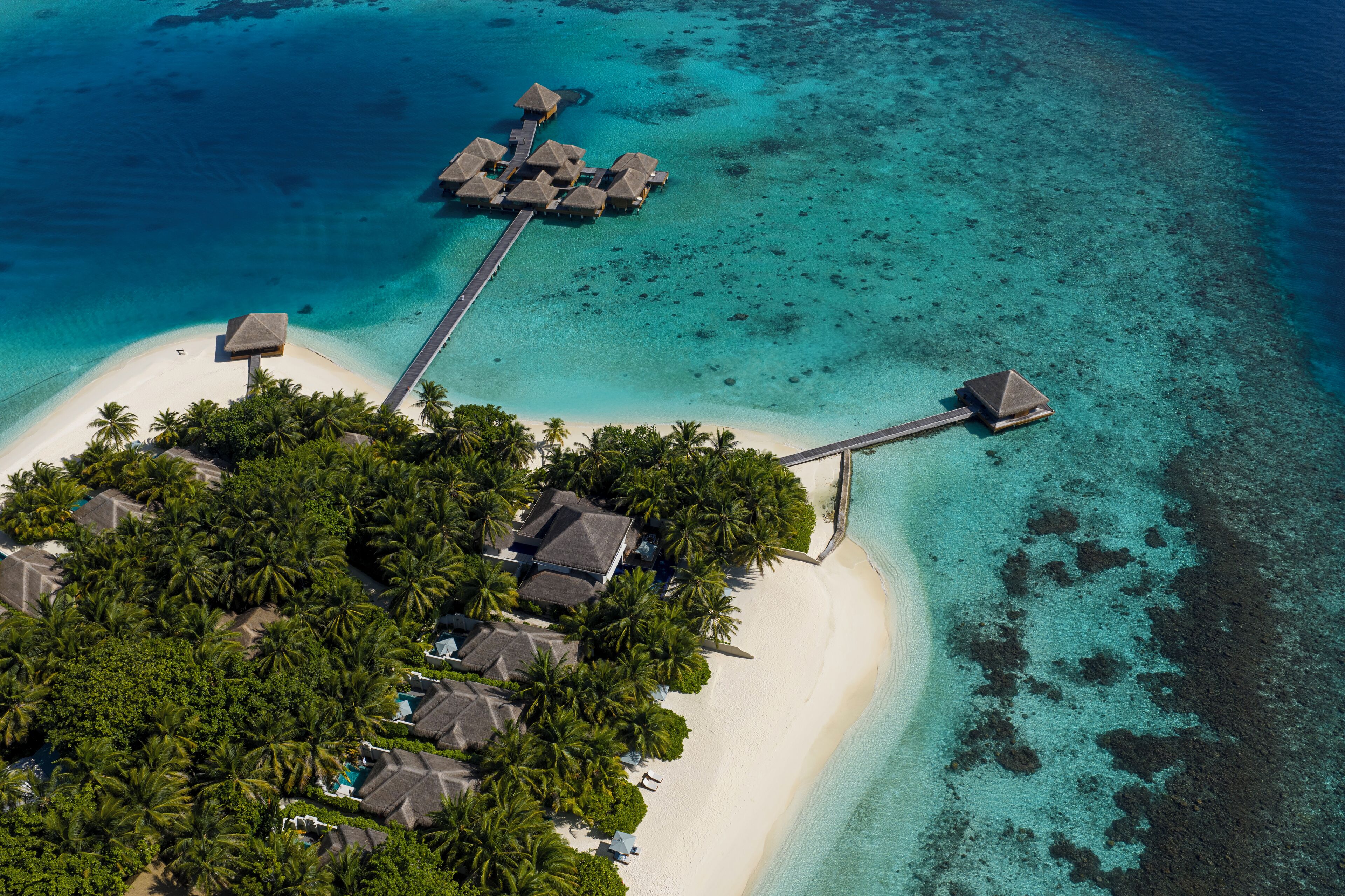 Vista aérea de un resort tropical con bungalows sobre el agua conectados por pasarelas, una playa de arena y abundante vegetación.