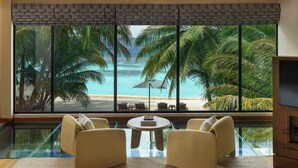 Een kamer met een glazen vloer, twee fauteuils, een kleine tafel en een groot raam met uitzicht op een strand en palmbomen.