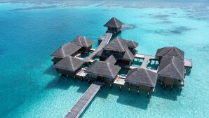 Een resort met bungalows op palen, meerdere rieten hutten verbonden door loopbruggen boven helder turquoise water.