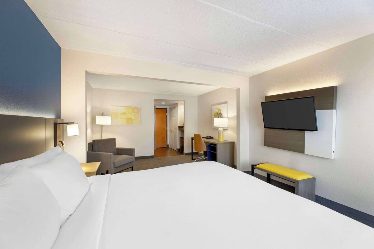 A hotel room with a large bed, a desk with a chair, a TV mounted on the wall, and a small bench.