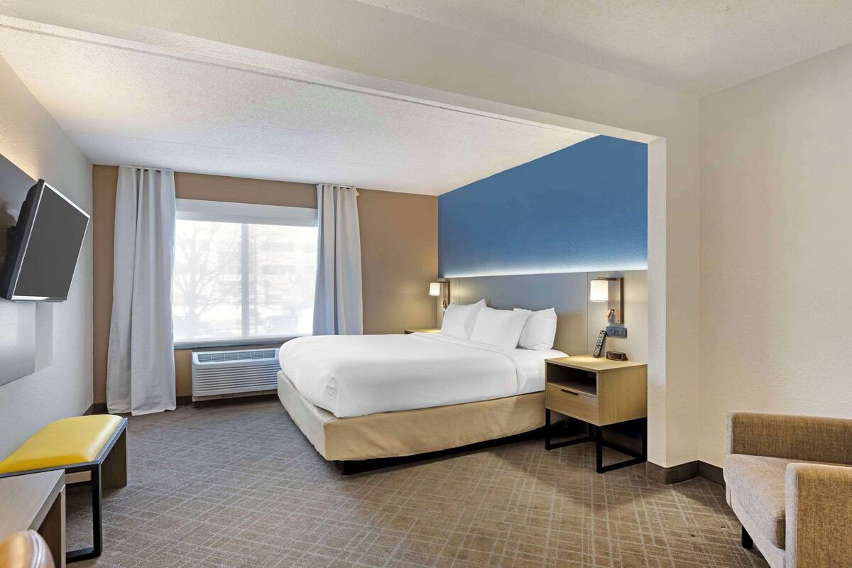 A hotel room with a bed, bedside table, lamp, TV, and a window with curtains.