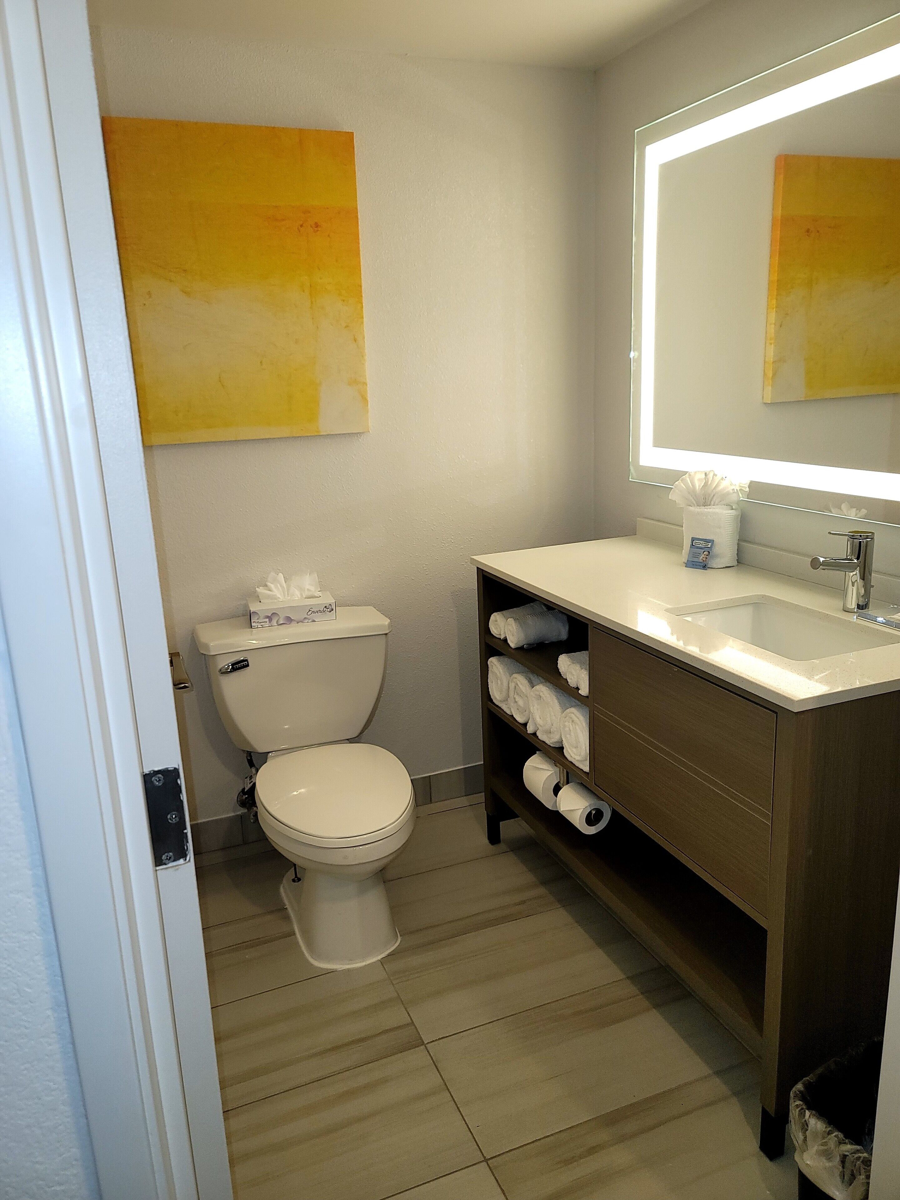 Un baño con inodoro, lavamanos y espejo. Hay toallas y papel higiénico en la encimera.
