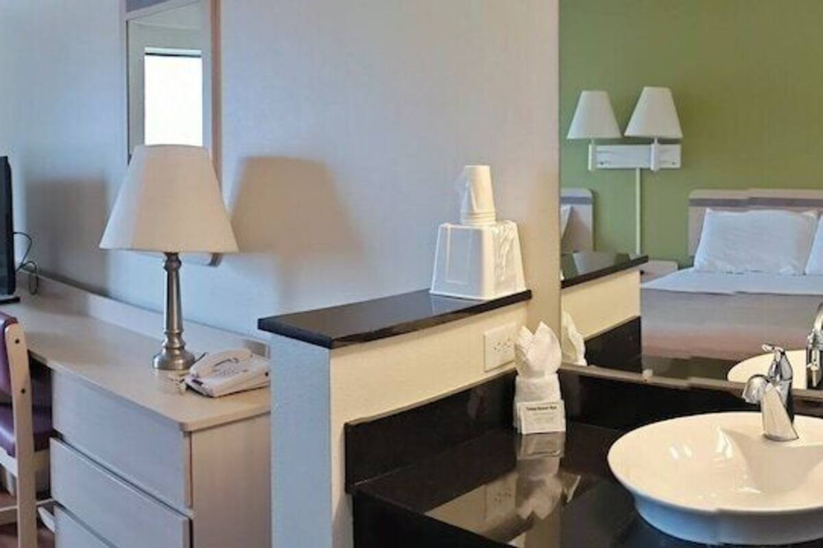 A hotel room with two beds, a desk with a computer, a bathroom with a sink and mirror, and a window with curtains.