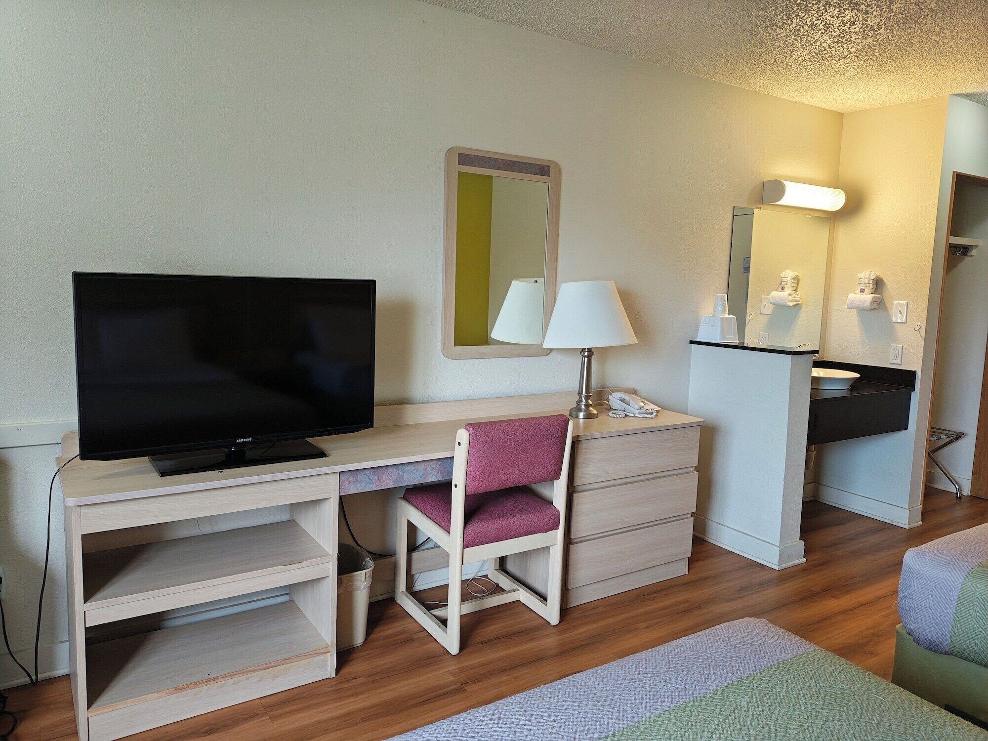 A hotel room with a TV, a desk with a lamp, a chair, a dresser, and a mirror.