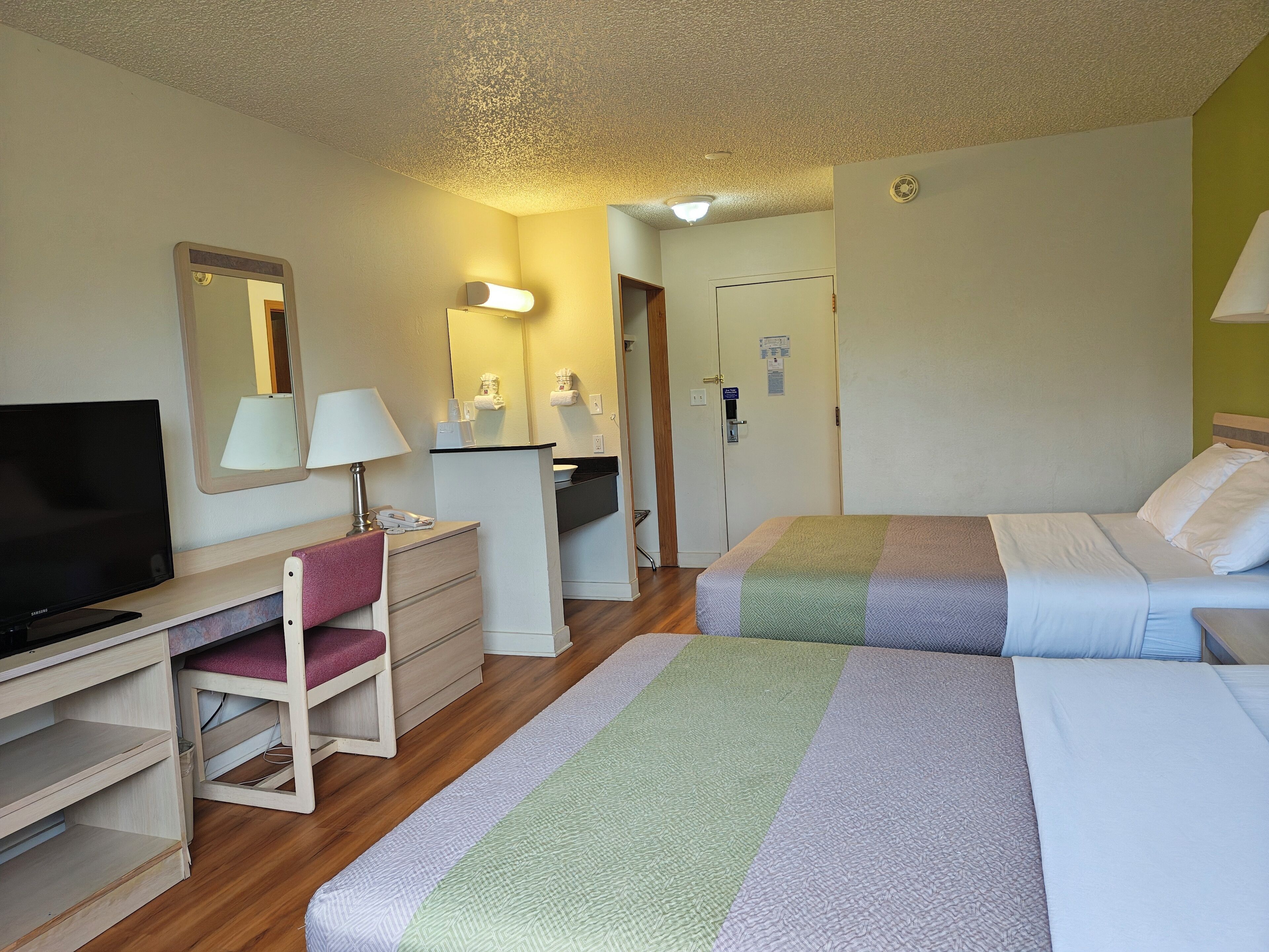 A hotel room with a bed, a desk with a TV, a chair, a mirror, and a door.