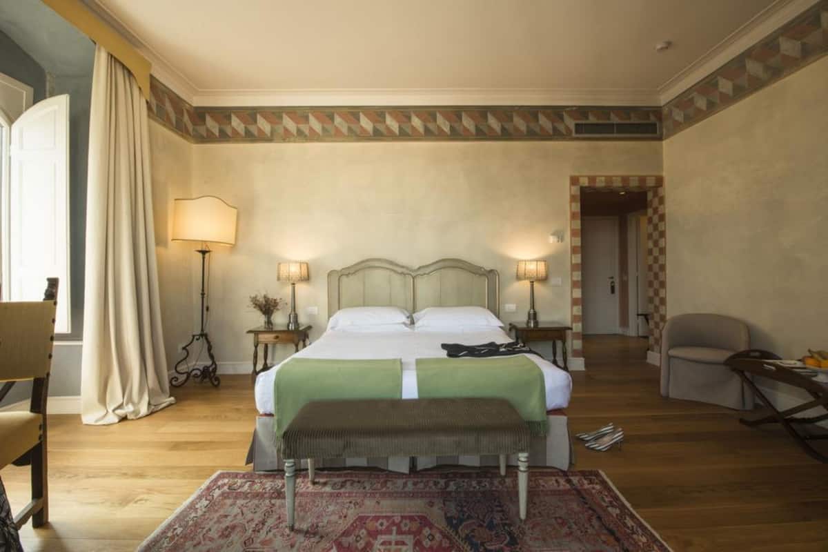 Une chambre d’hôtel avec un grand lit, deux lampes de chevet, un banc, une chaise et un tapis à motifs.