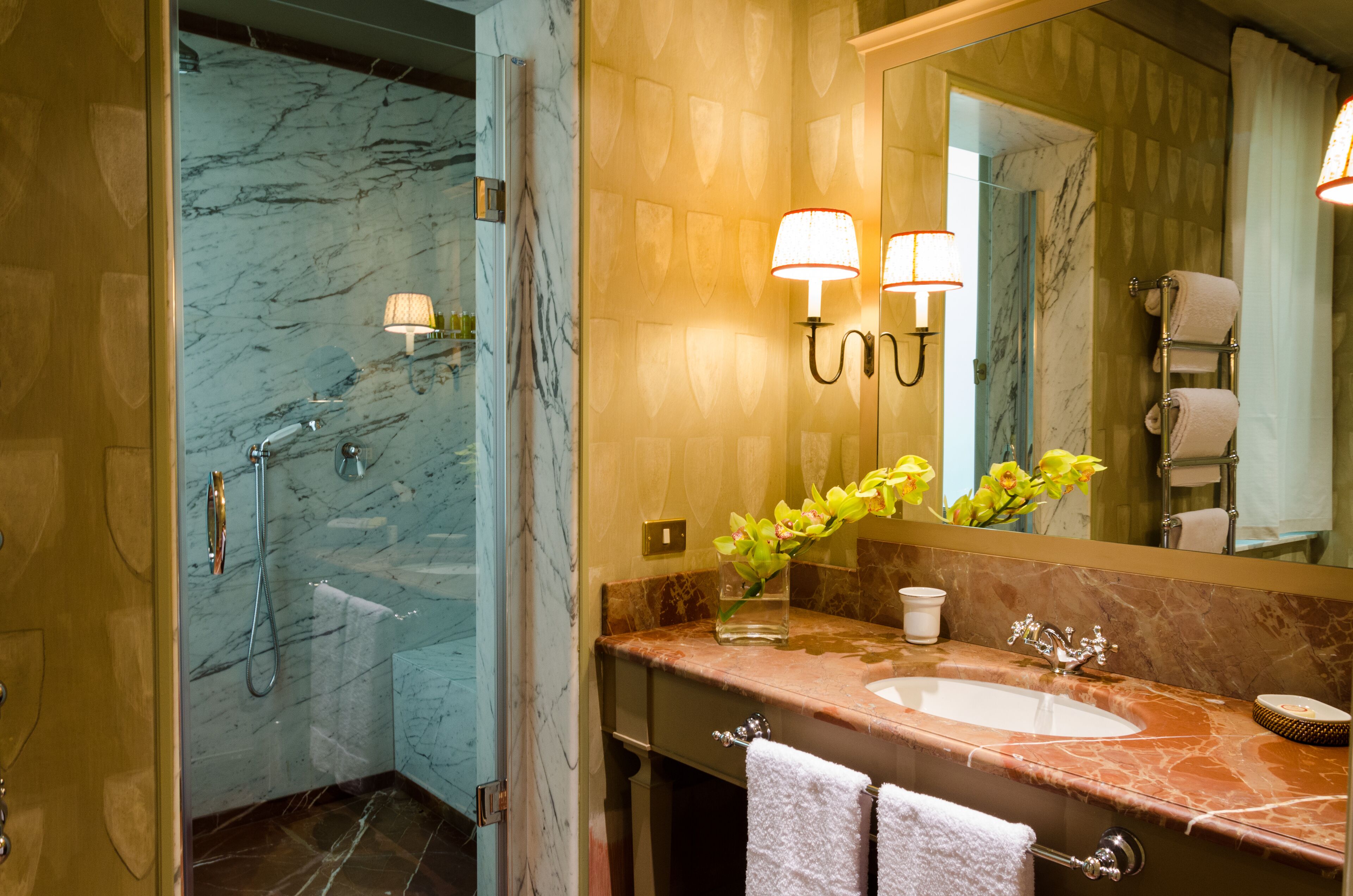A bathroom with a marble shower, a sink with a vase of flowers, and two wall-mounted lamps.