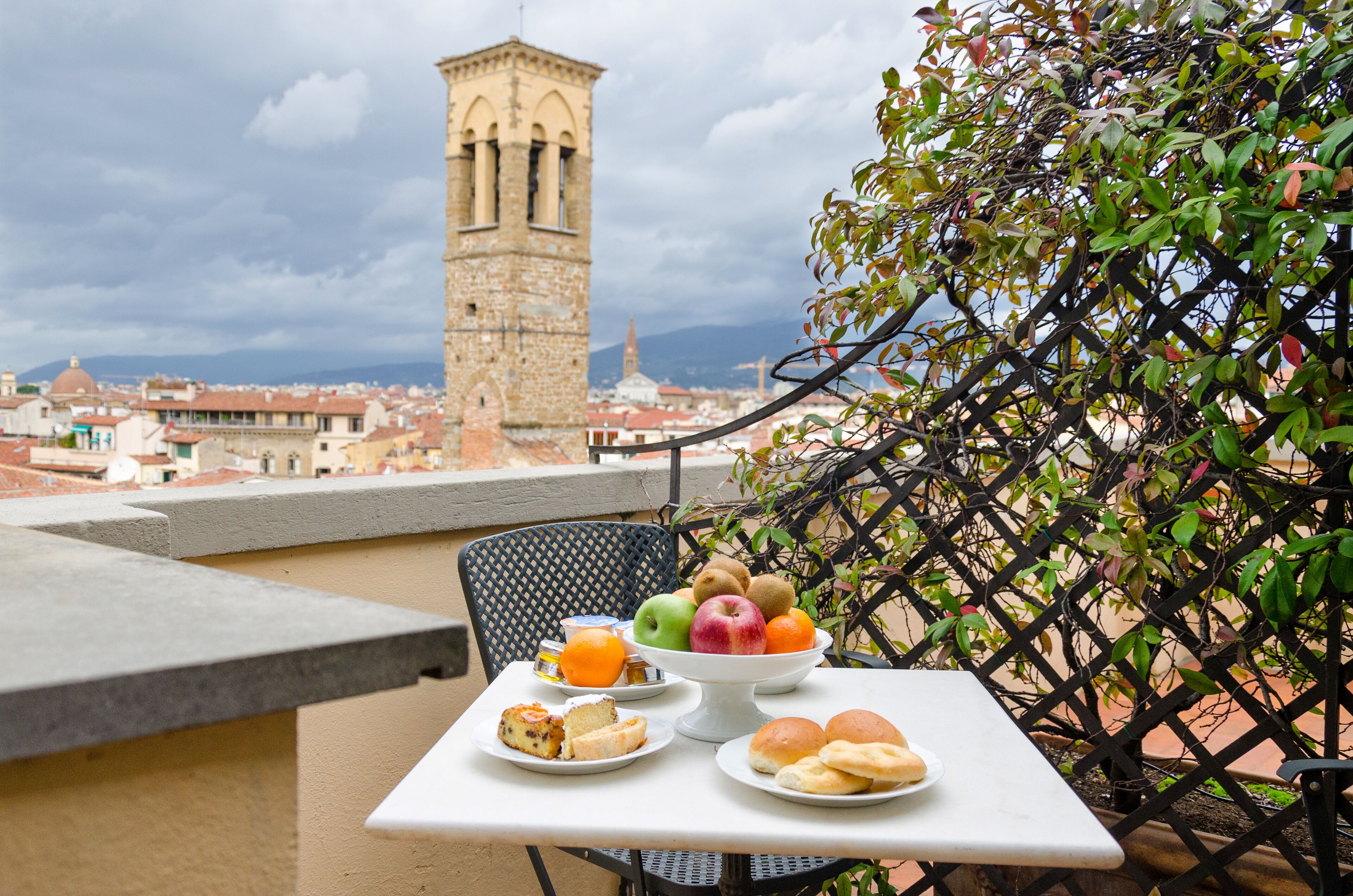 A table with a fruit bowl, bread, and a drink on a balcony overlooking a cityscape with a tower.