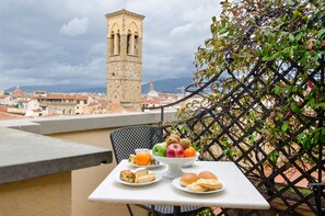 Une table avec un panier de fruits, du pain et une boisson, placée sur un balcon donnant sur un paysage urbain avec une tour.