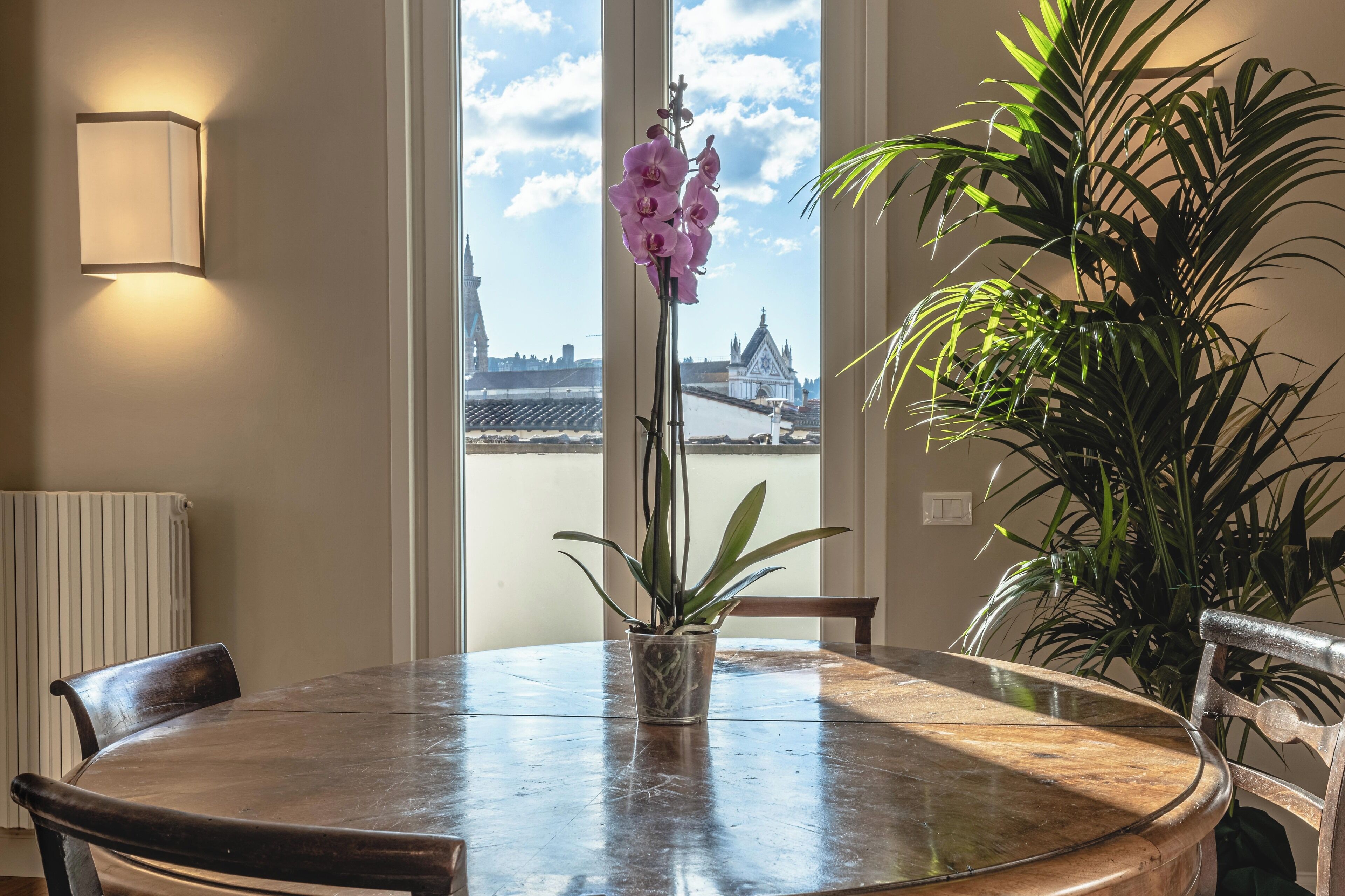 A round dining table with chairs, a potted plant, and a view of a cityscape through large windows.