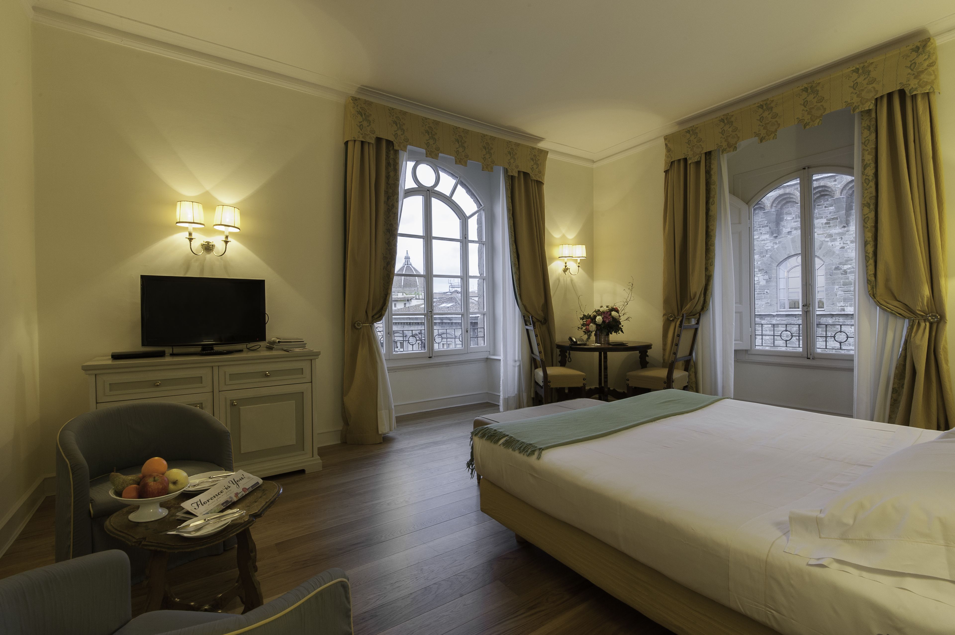 Habitación de hotel con cama, escritorio con televisor, silla, mesita con frutero y dos ventanas con cortinas.