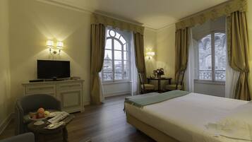 Habitación de hotel con cama, escritorio con televisor, silla, mesita con frutero y dos ventanas con cortinas.