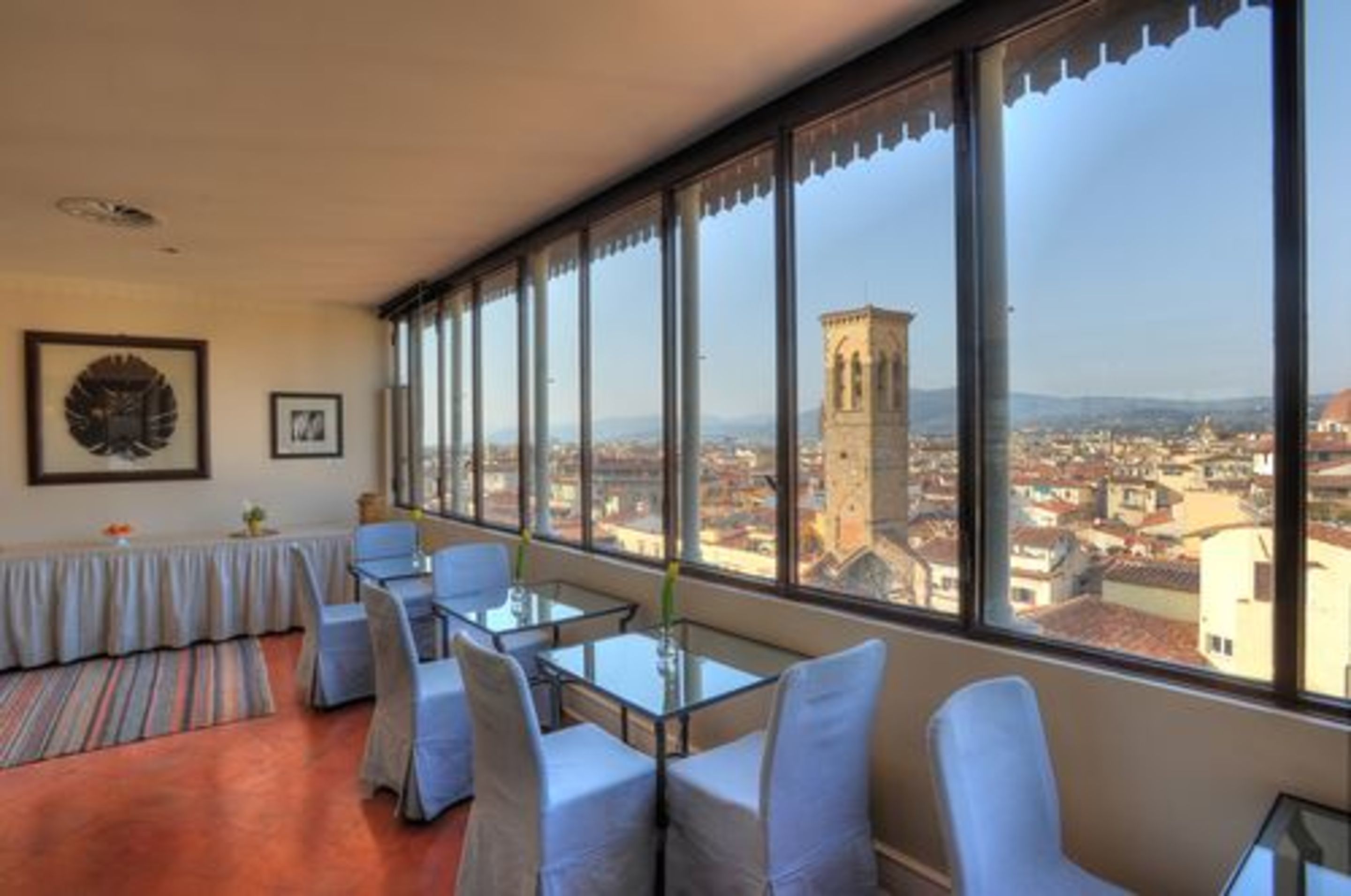 A hotel room with large windows offering a city view, a dining area with tables and chairs, and framed artwork on the wall.