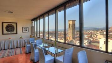 A hotel room with large windows offering a city view, a dining area with tables and chairs, and framed artwork on the wall.