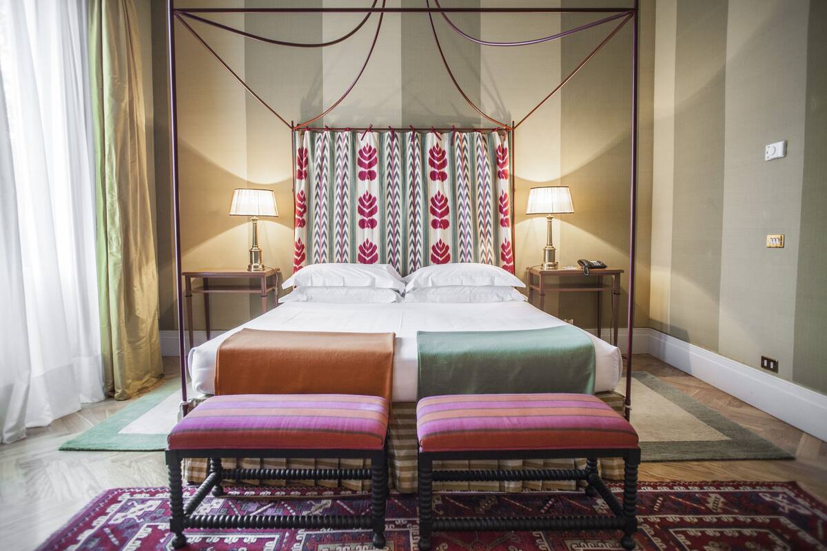 Un lit double à baldaquin, deux tables de chevet avec des lampes, une paire d’ottomans et un tapis à motifs.