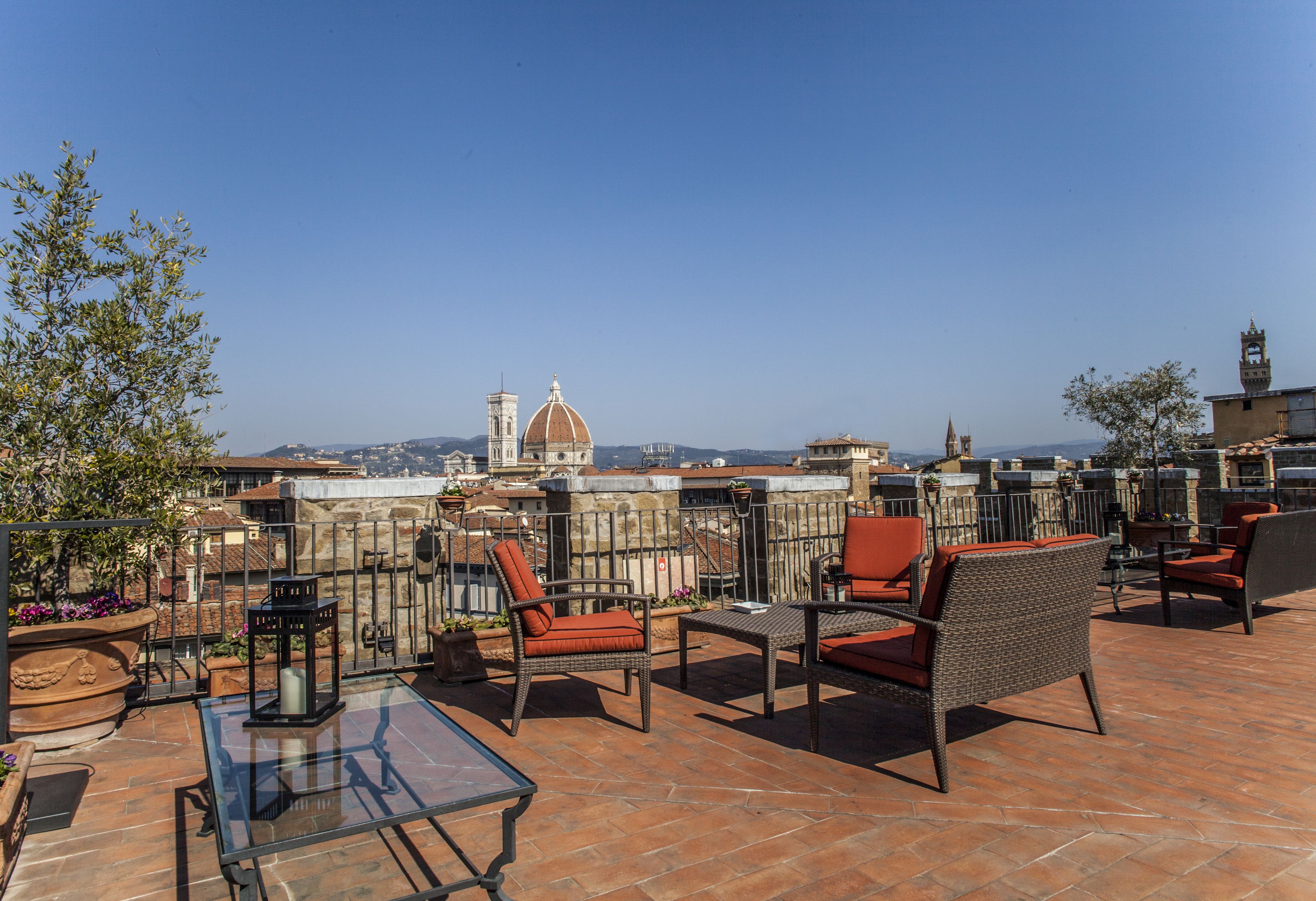 Una terraza en la azotea con mobiliario de mimbre, una mesa de vidrio y vistas a un edificio histórico con cúpula y campanario.