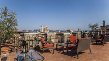 Una terraza en la azotea con mobiliario de mimbre, una mesa de vidrio y vistas a un edificio histórico con cúpula y campanario.