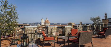 Una terraza en la azotea con mobiliario de mimbre, una mesa de vidrio y vistas a un edificio histórico con cúpula y campanario.
