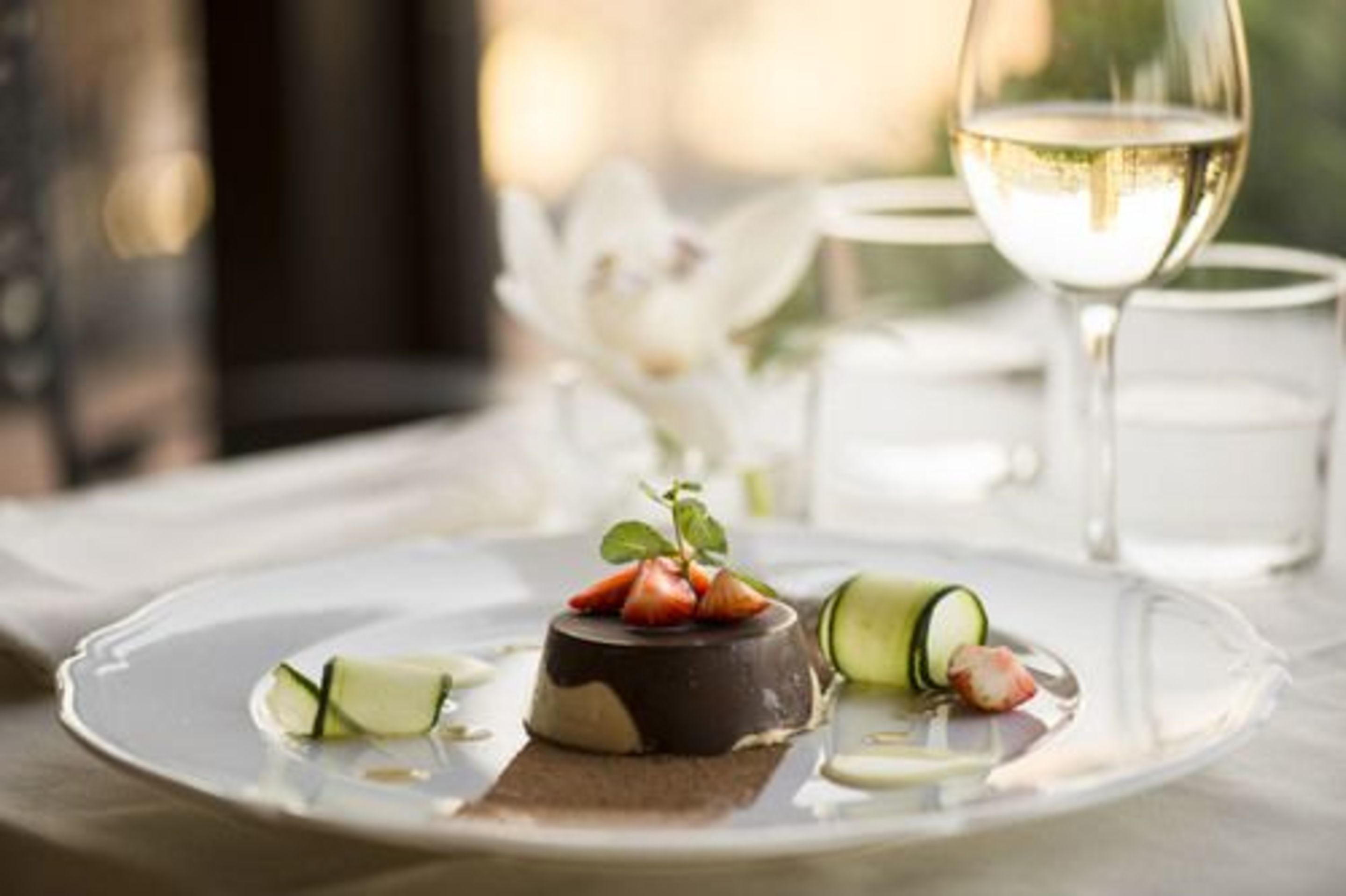 A dessert plate with a chocolate dome, strawberries, and cucumber slices, accompanied by a glass of white wine.
