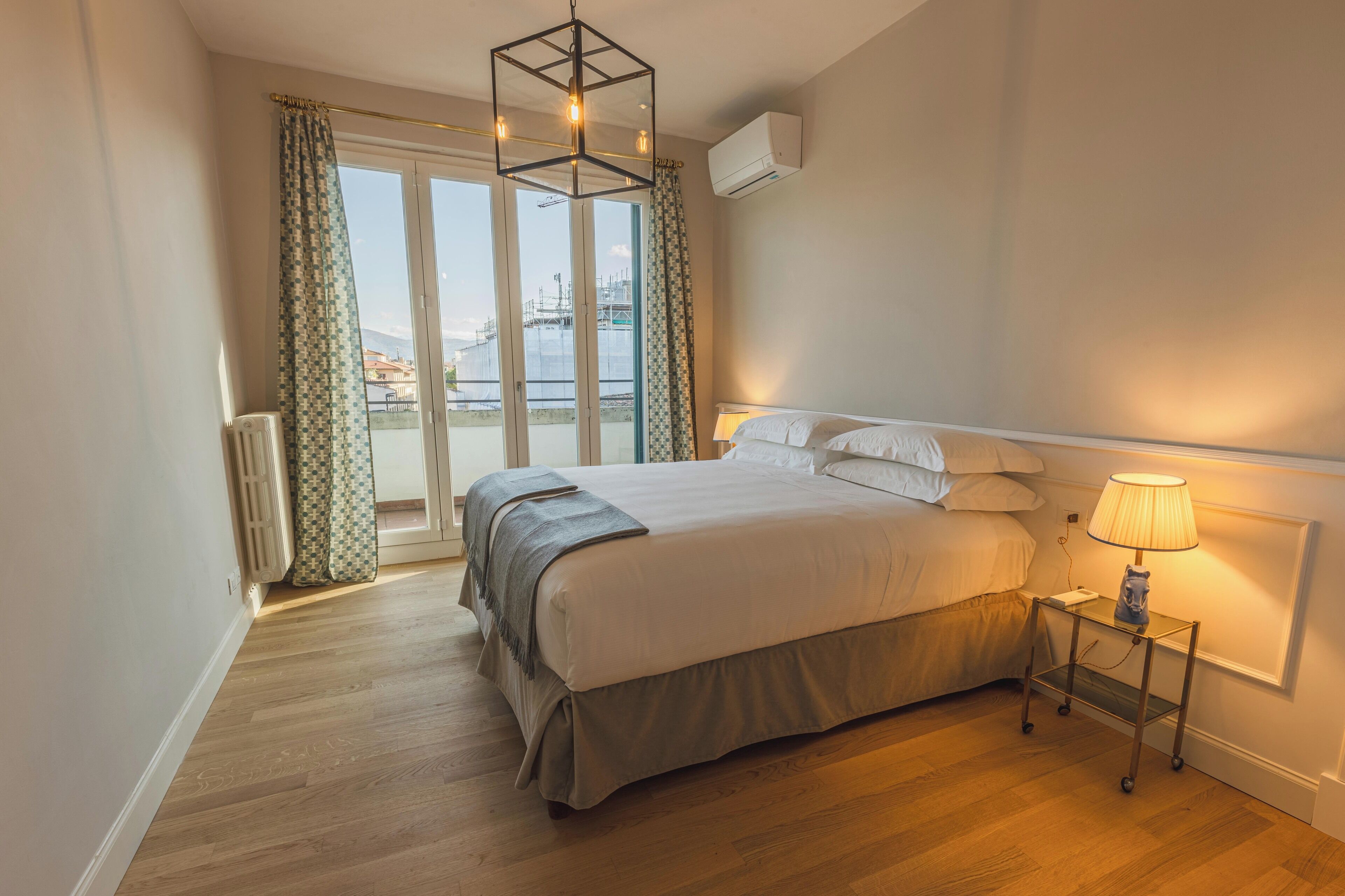 Quarto com cama grande, mesa de cabeceira com abajur, radiador e vista para a cidade pela janela.