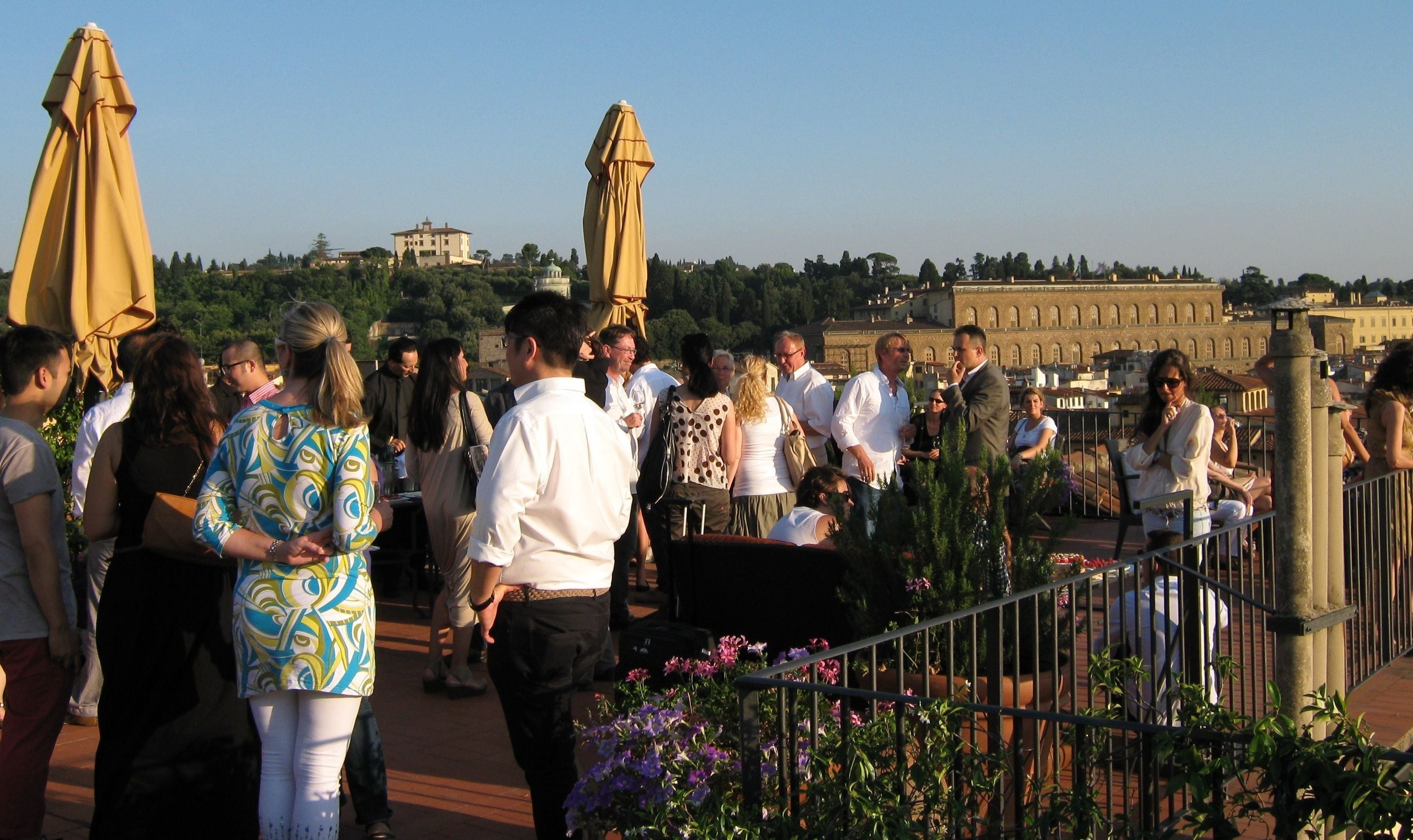 A rooftop terrace with people gathered, outdoor seating, and a view of historic buildings in the background.