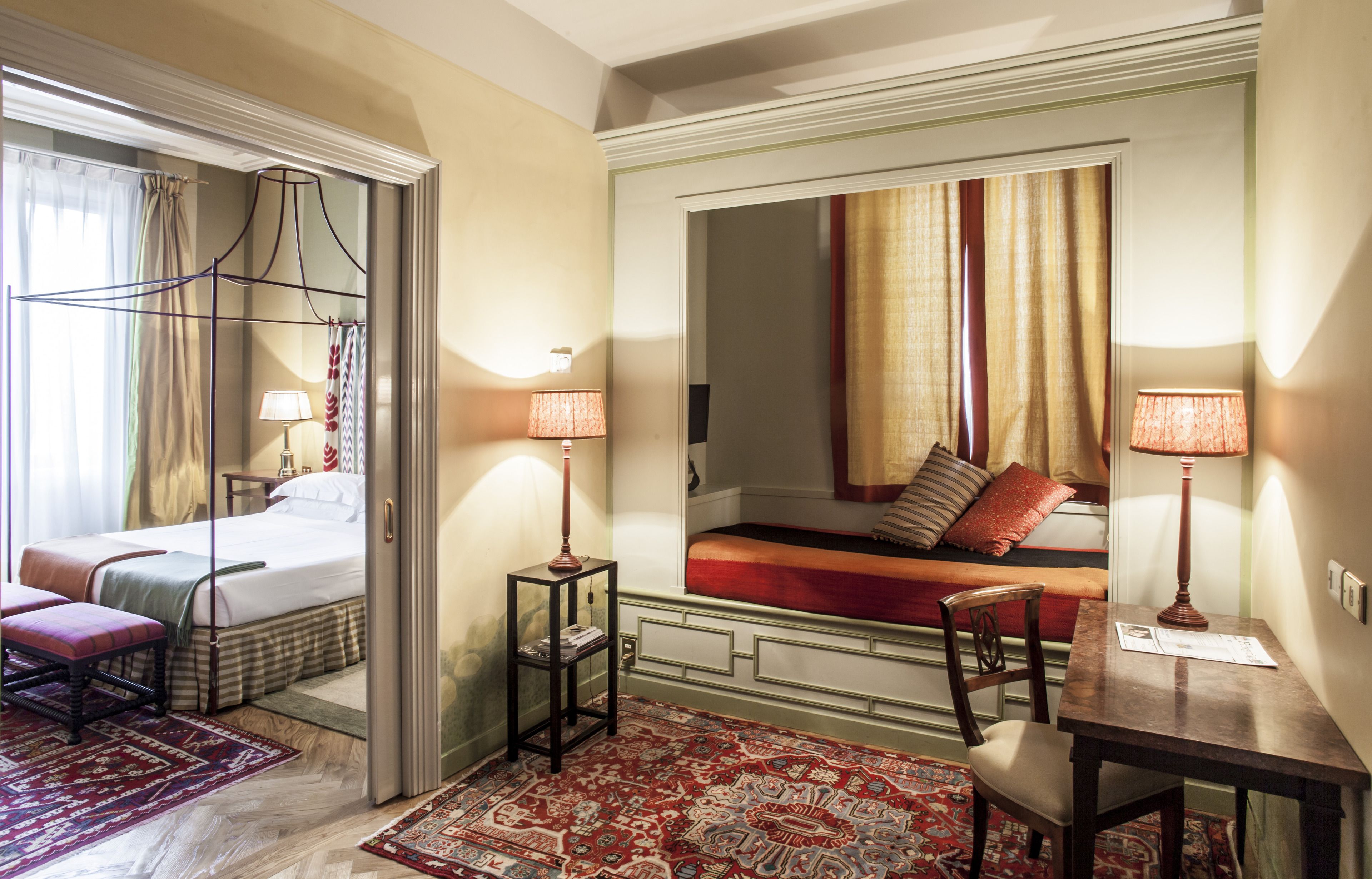 A hotel room with a bed, a sitting area with a sofa, a table, and a chair. There are lamps on the table and a mirror on the wall.
