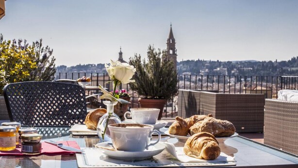 Une table dressée avec des croissants, des tasses de café et un vase rempli de fleurs, sur un balcon donnant sur une vue panoramique de la ville.
