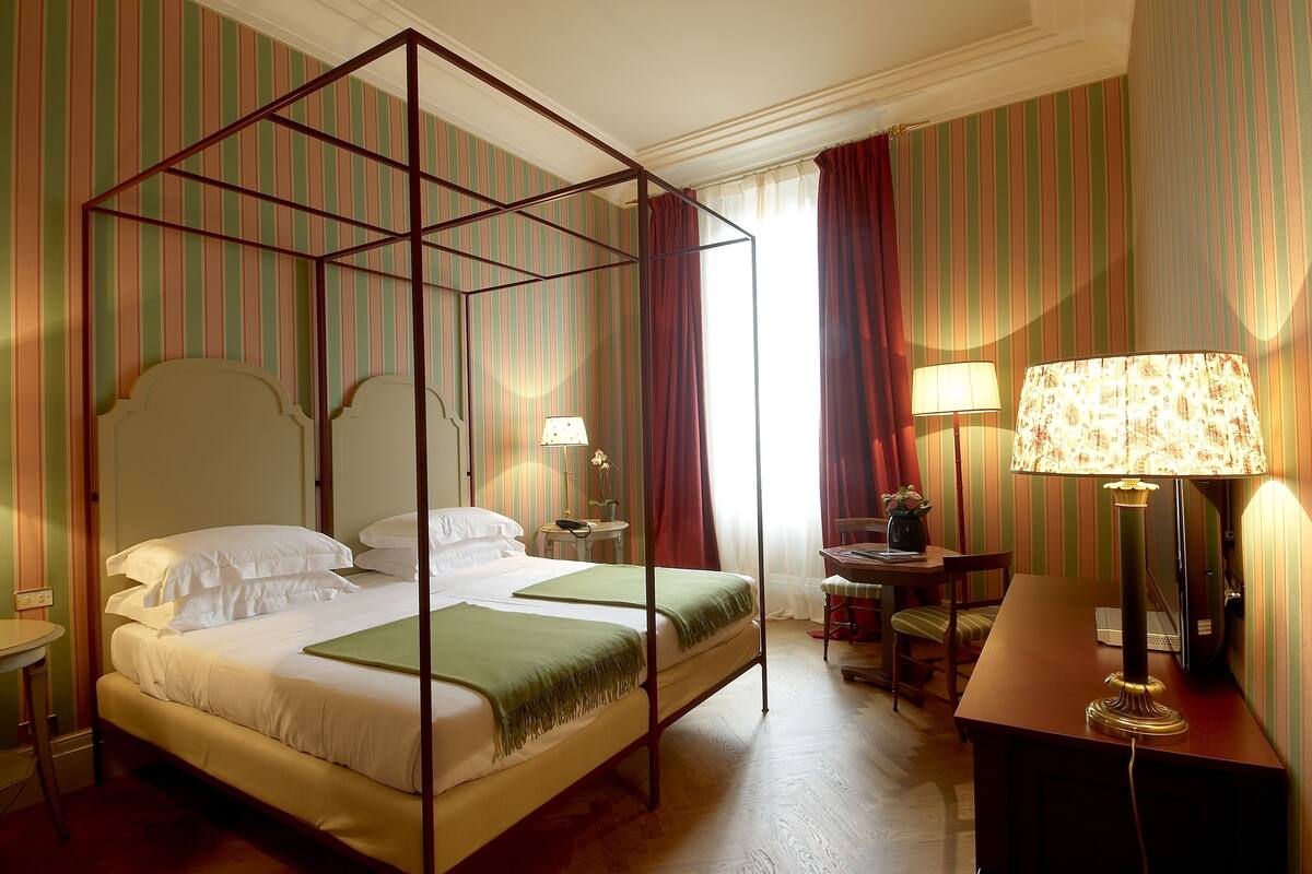 Un lit à baldaquin avec un plaid vert, un bureau avec une lampe et une chaise, dans une chambre d’hôtel aux murs recouverts d’un papier peint à rayures.