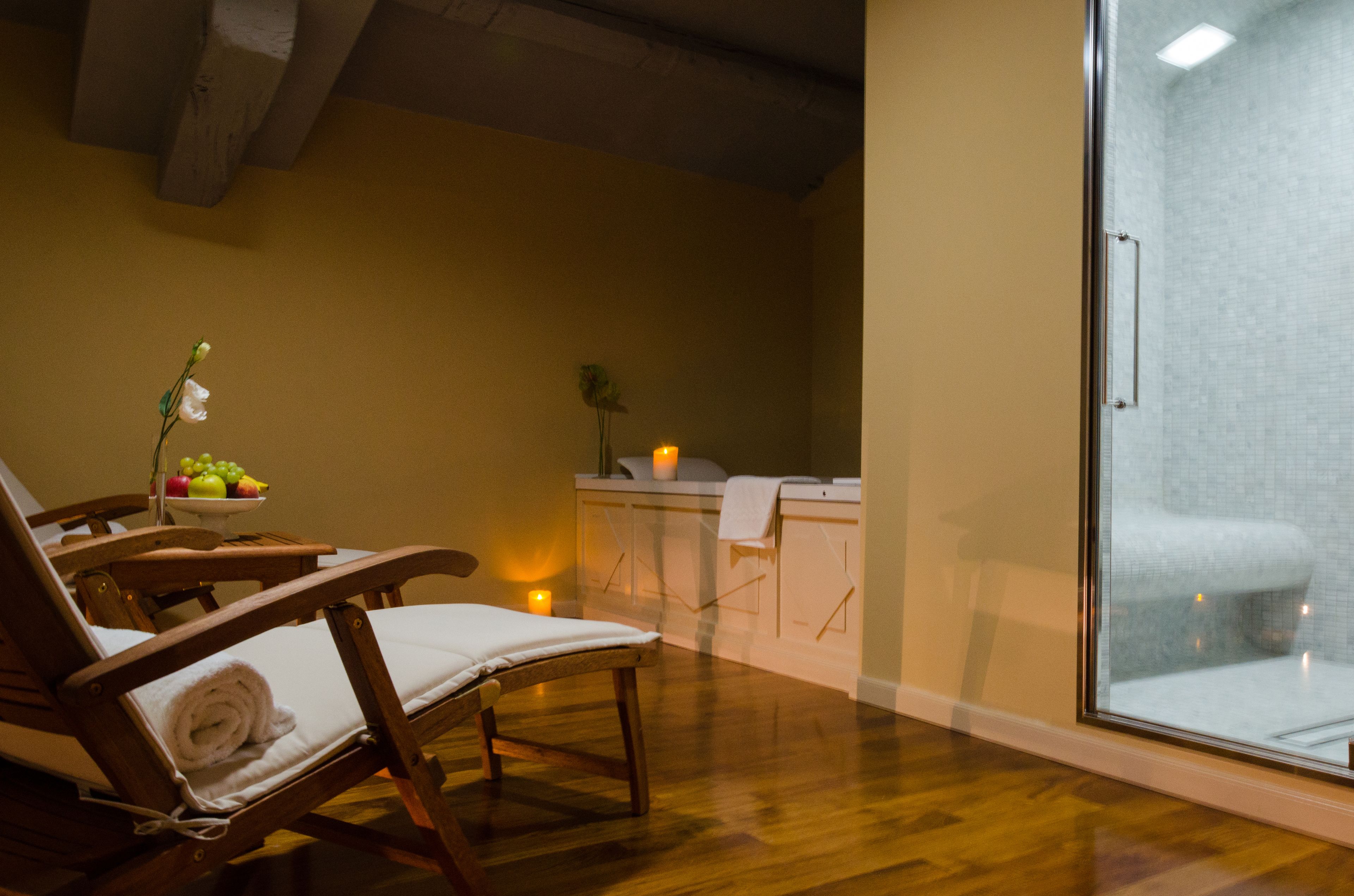 A spa room with wooden chairs, a small table with fruit, a massage table, and a glass shower enclosure.